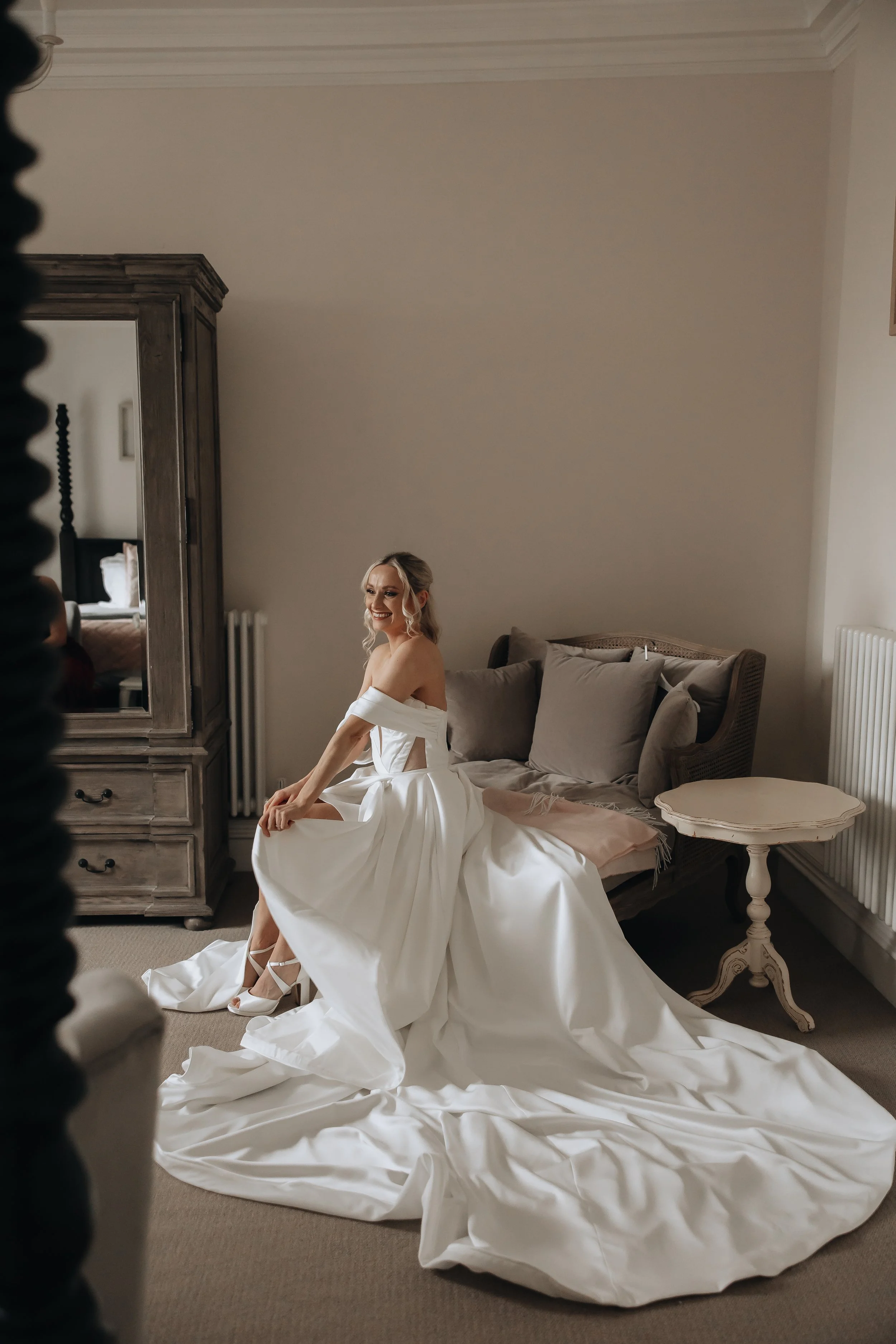 A woman in a white wedding gown sitting on a sofa, smiling, in a softly lit room with neutral-colored furniture.